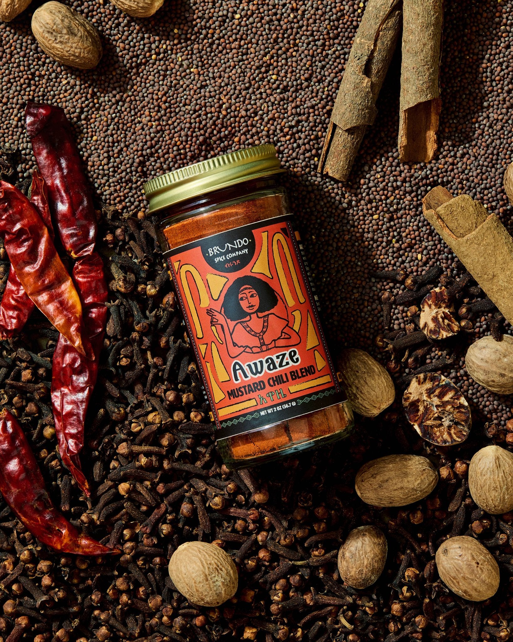 Brundo Spice Company Awaze Mustard Chili Blend jar surrounded by dried chili peppers, mustard seeds, cloves, nutmeg, and cinnamon bark.