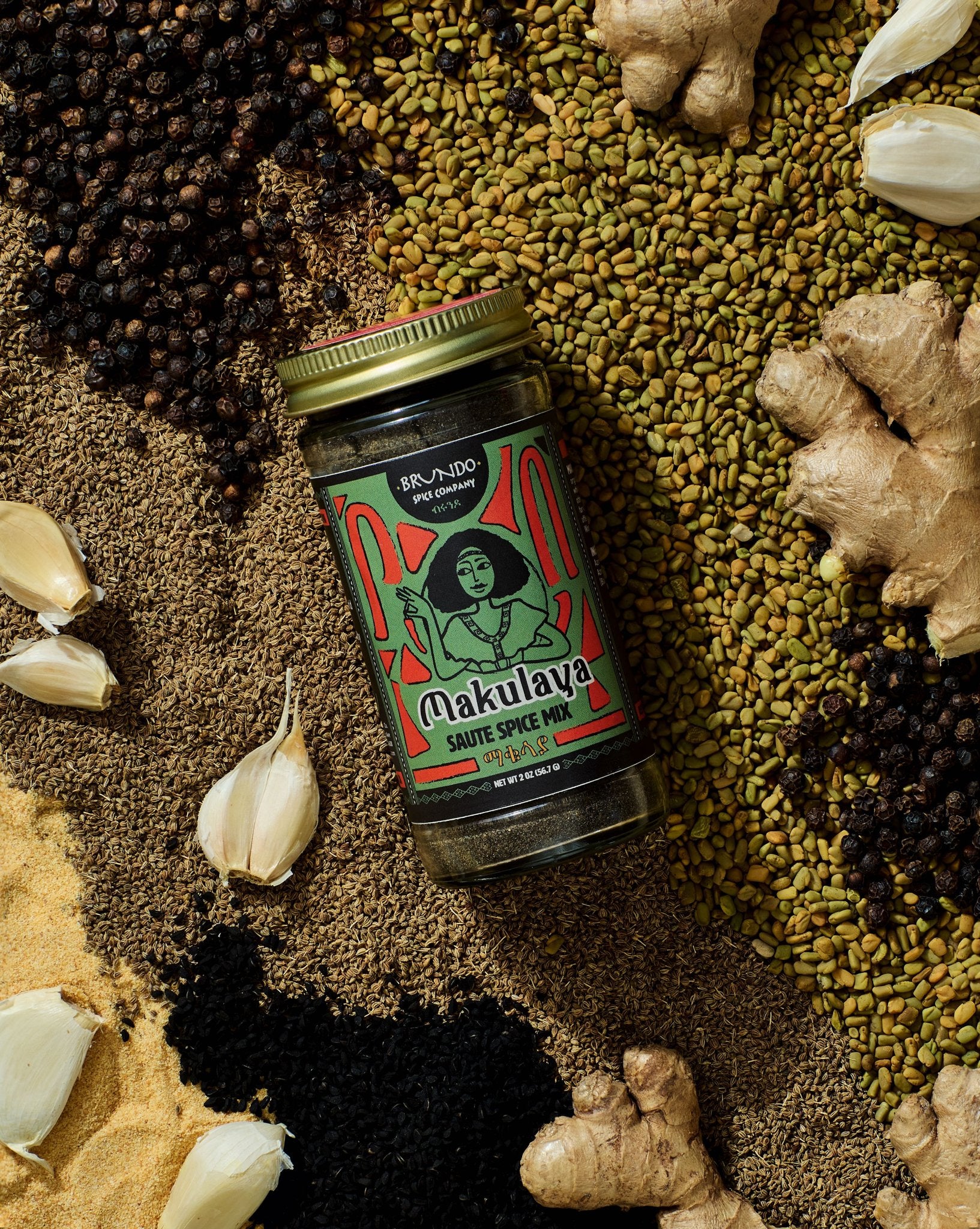 Brundo Spice Company Makulaya Saute Spice Mix jar displayed on a bed of whole and ground spices including garlic, black seeds, and ginger.