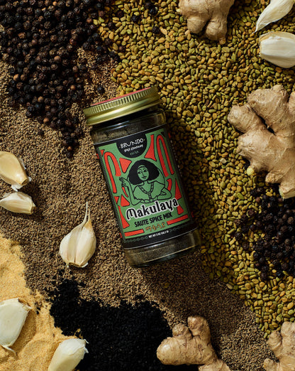Brundo Spice Company Makulaya Saute Spice Mix jar displayed on a bed of whole and ground spices including garlic, black seeds, and ginger.