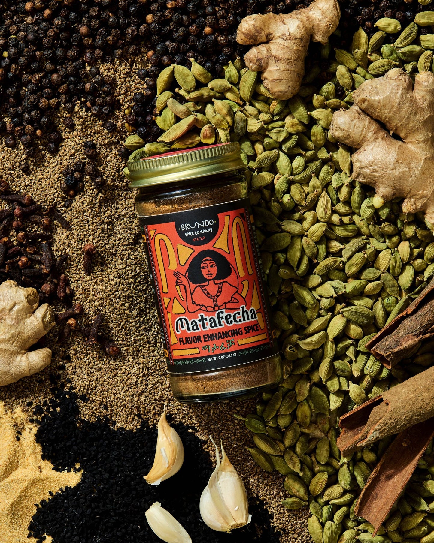 Brundo Spice Company Matafecha Flavor Enhancing Spice jar surrounded by green cardamom, ajwain, ginger, cinnamon bark, and garlic.