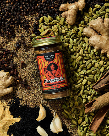Brundo Spice Company Matafecha Flavor Enhancing Spice jar surrounded by green cardamom, ajwain, ginger, cinnamon bark, and garlic.
